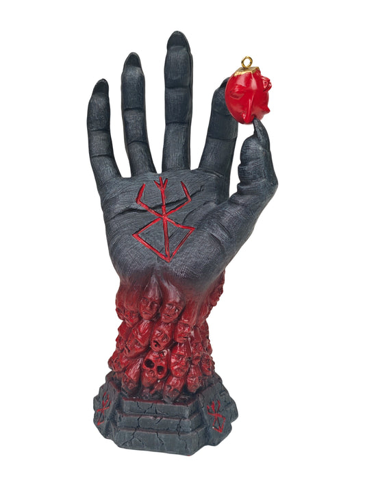 A gothic horror ornament shaped like a severed hand holding a small red skull, with a red skull-filled base and red symbols on a white background.