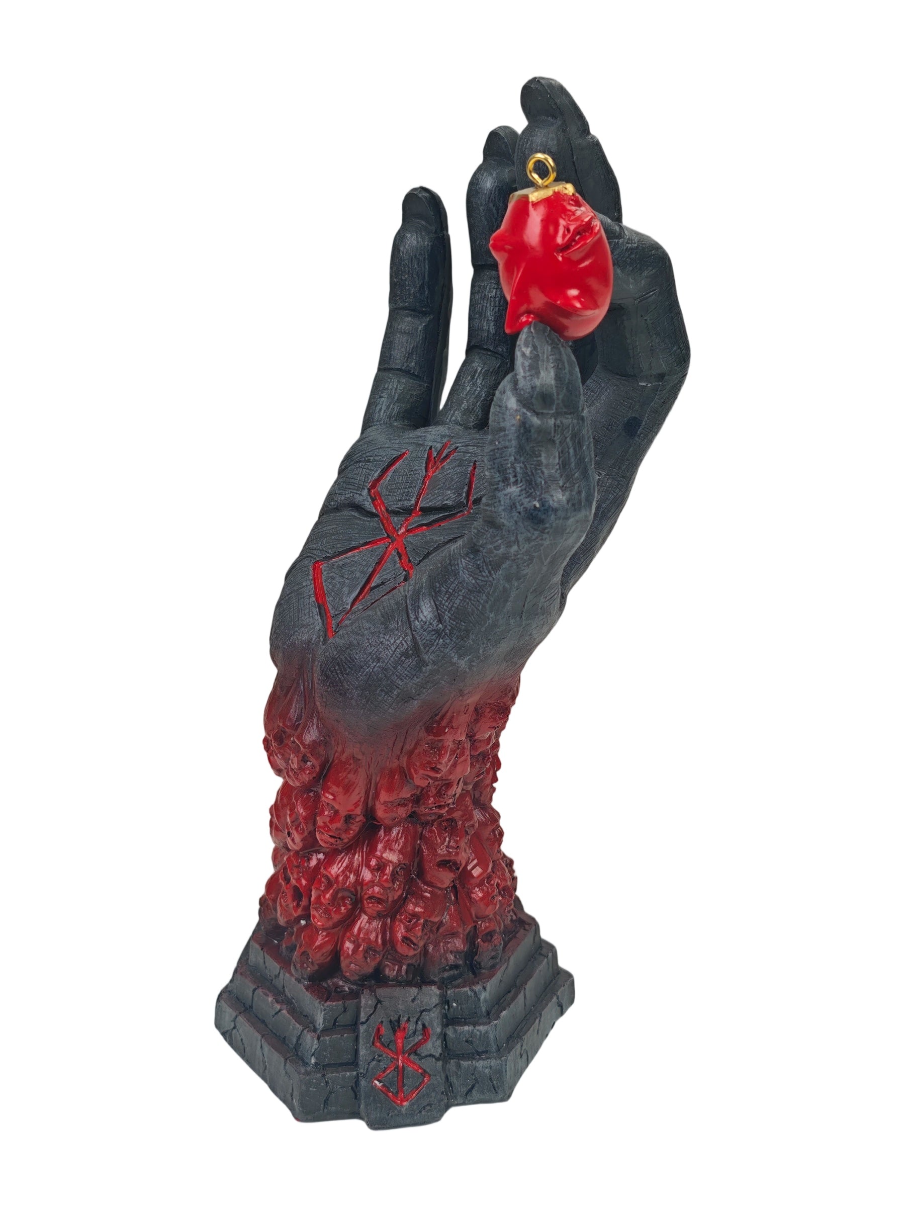 A gothic horror-themed sculpture of a skeletal hand rising from a stone-like base, with red carved markings on the palm and a bright red heart-shaped pendant held in the fingers against a plain white background.