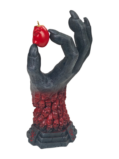 A gothic-style ornament featuring a dark, skeletal hand holding a small red apple with a gold loop, with the hand transitioning into a base of red skulls on a white background.